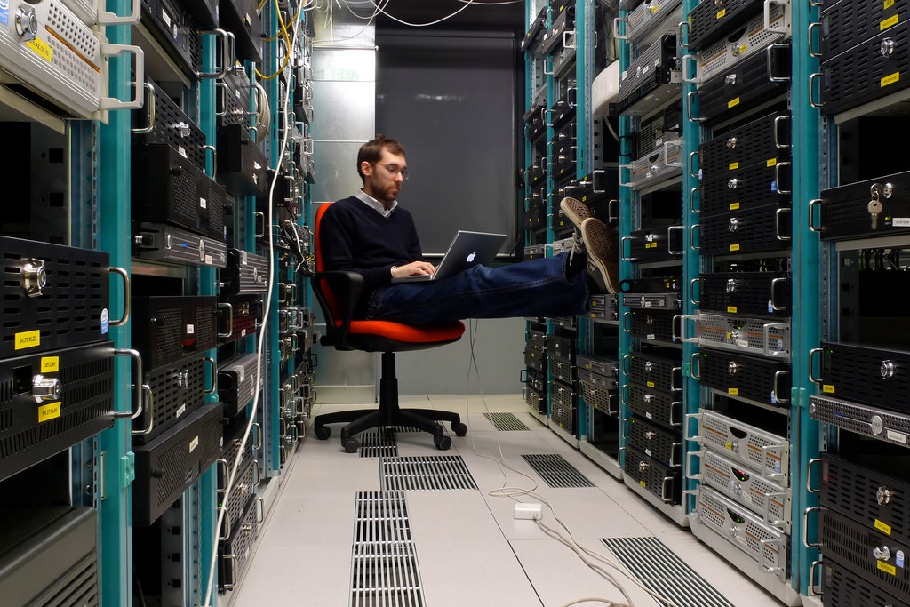 A man in glasses and a navy jumper sitting on an orange office chair with his feet up, working on a laptop in a server room between tall racks of equipment