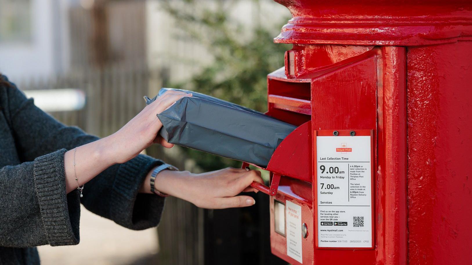 A person in a grey knitted cardigan posting a grey parcel into a red Royal Mail pillar box, with the last collection times visible on the box