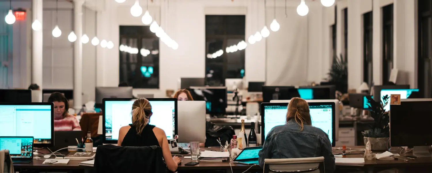 People working at computers in a bright open-plan office with hanging bulb lights