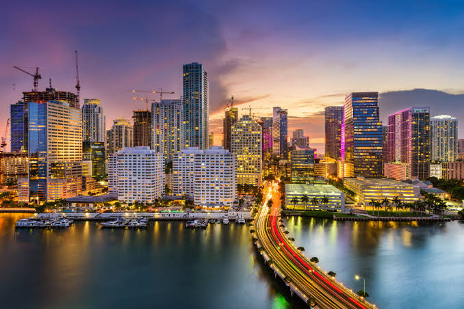 Miami skyline at dusk with lit high-rise towers, boats in a marina and light trails on a causeway over the water